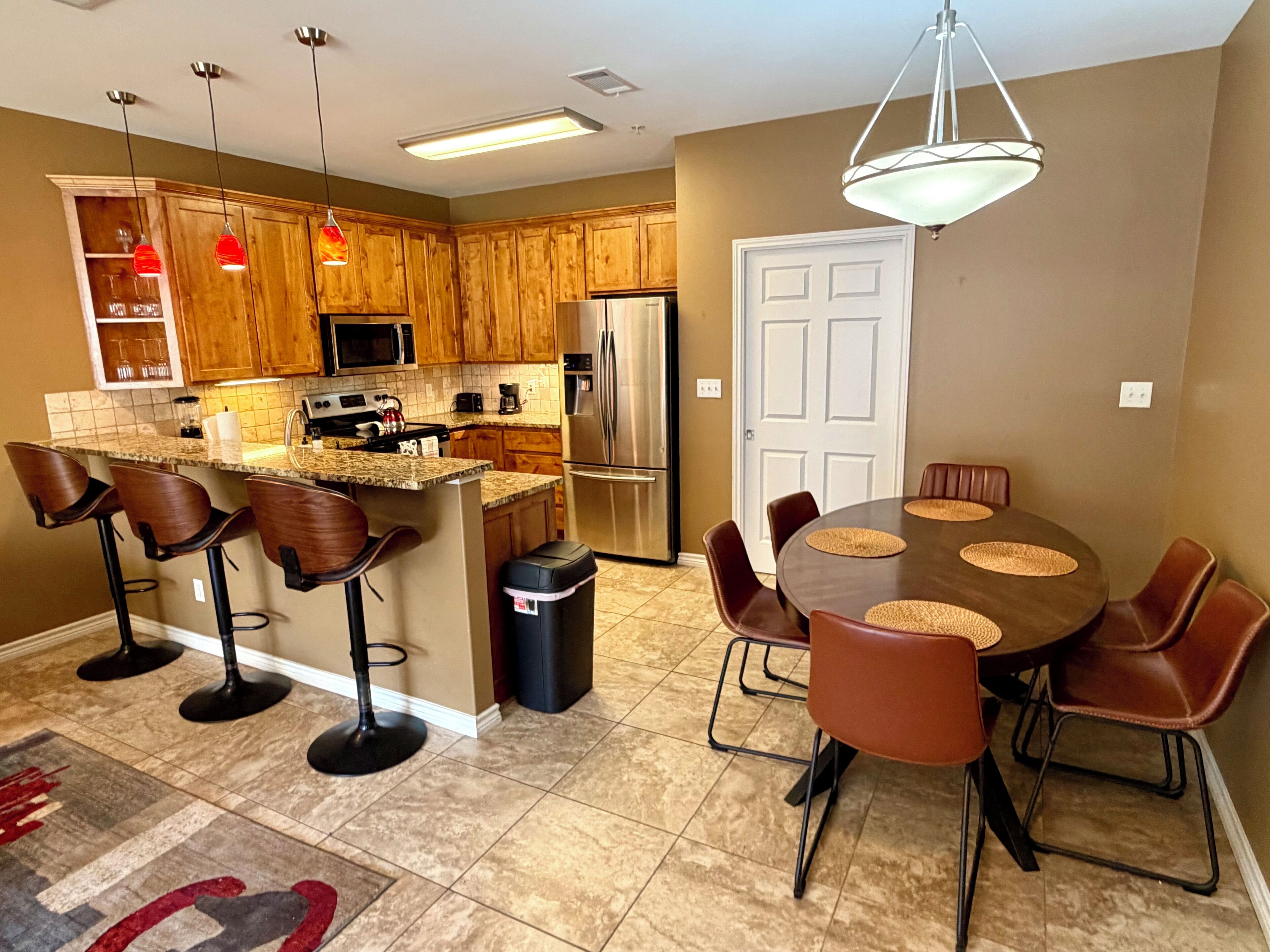 A modern kitchen with wooden cabinets, a central island with bar stools, a dining table with chairs, and a refrigerator.