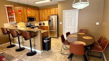 A modern kitchen with wooden cabinets, a central island with bar stools, a dining table with chairs, and a refrigerator.