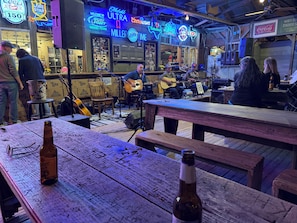 Un bar avec des enseignes au néon, une scène où des musiciens jouent, et des tables en bois avec des bouteilles.