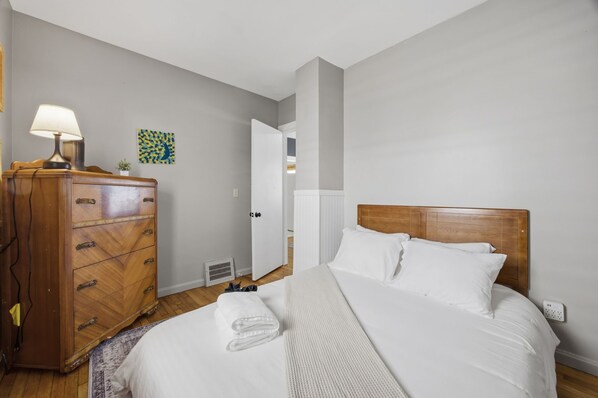 A bedroom with a wooden dresser, a bed with white linens, a nightstand, and a wall-mounted lamp. - 3 Private Apts in Tremont - Perfect for Groups (Cleveland)