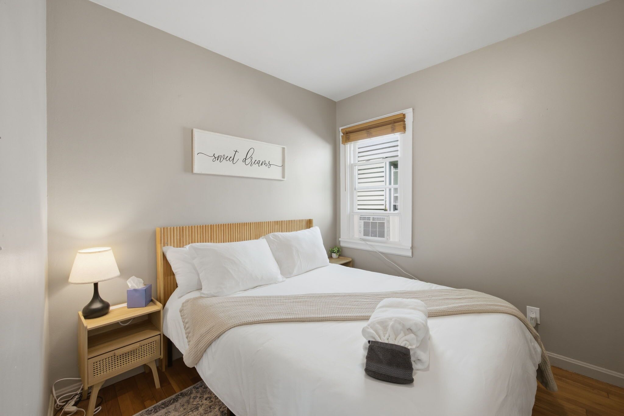 A neatly made bed with a headboard, a bedside lamp, a small table with a lamp, and a window with blinds.