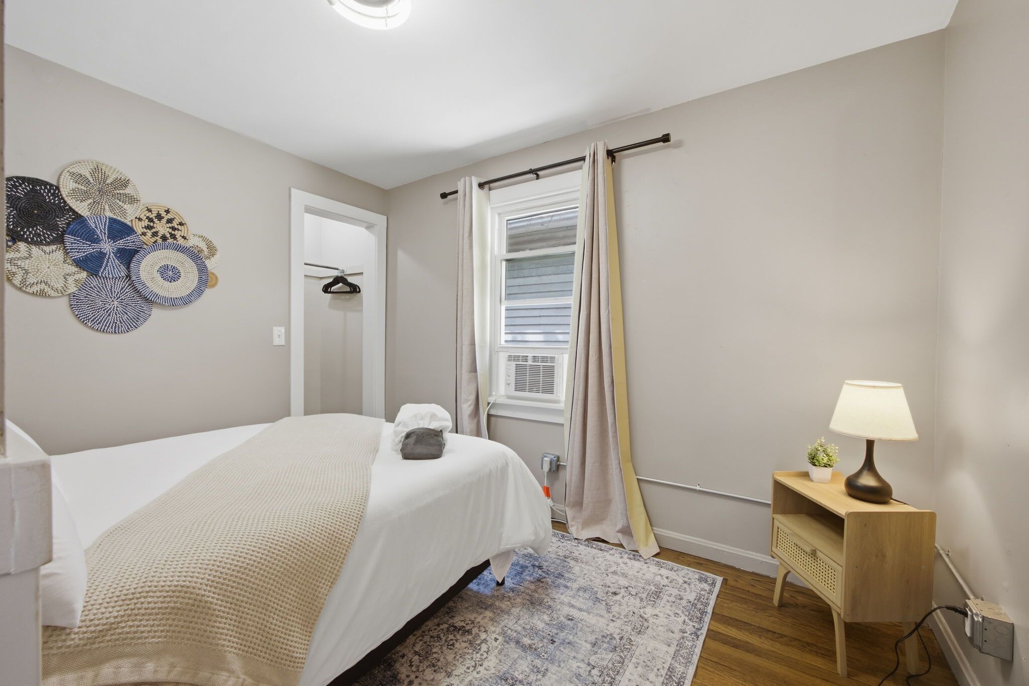 Un dormitorio con cama, mesita de noche con lámpara, estante en la pared, ventana con cortinas y decoración mural.