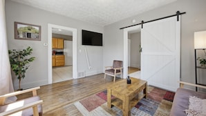 Un salon doté d’un plancher en bois, d’un téléviseur fixé au mur, d’une porte coulissante de style rustique et d’une cuisine visible à travers une porte ouverte.