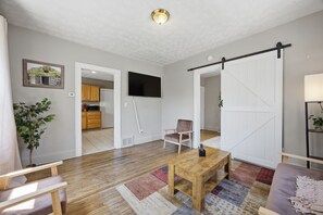 A living room with a wooden floor, a TV mounted on the wall, a sliding barn-style door, and a kitchen visible through an open door. - Bright Tremont 2BR - Near Downtown CLE & Clinic (Cleveland)