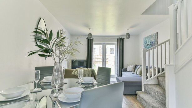 Een moderne eetruimte met een glazen tafel, witte borden en helder glaswerk. Op de achtergrond is een grijze bank en een trap met witte leuningen zichtbaar.