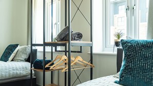 Een slaapkamer met een bed, een houten kledingrek, een radiator en een raam met uitzicht op gebouwen.