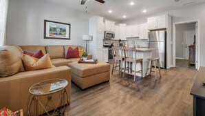 A modern living room with a sofa, ottoman, and coffee table, a dining area with wooden chairs, and a kitchen with white cabinets and stainless steel appliances. - Downtown Condo | 10 Min to FSU Stadium & Campus (Tallahassee)