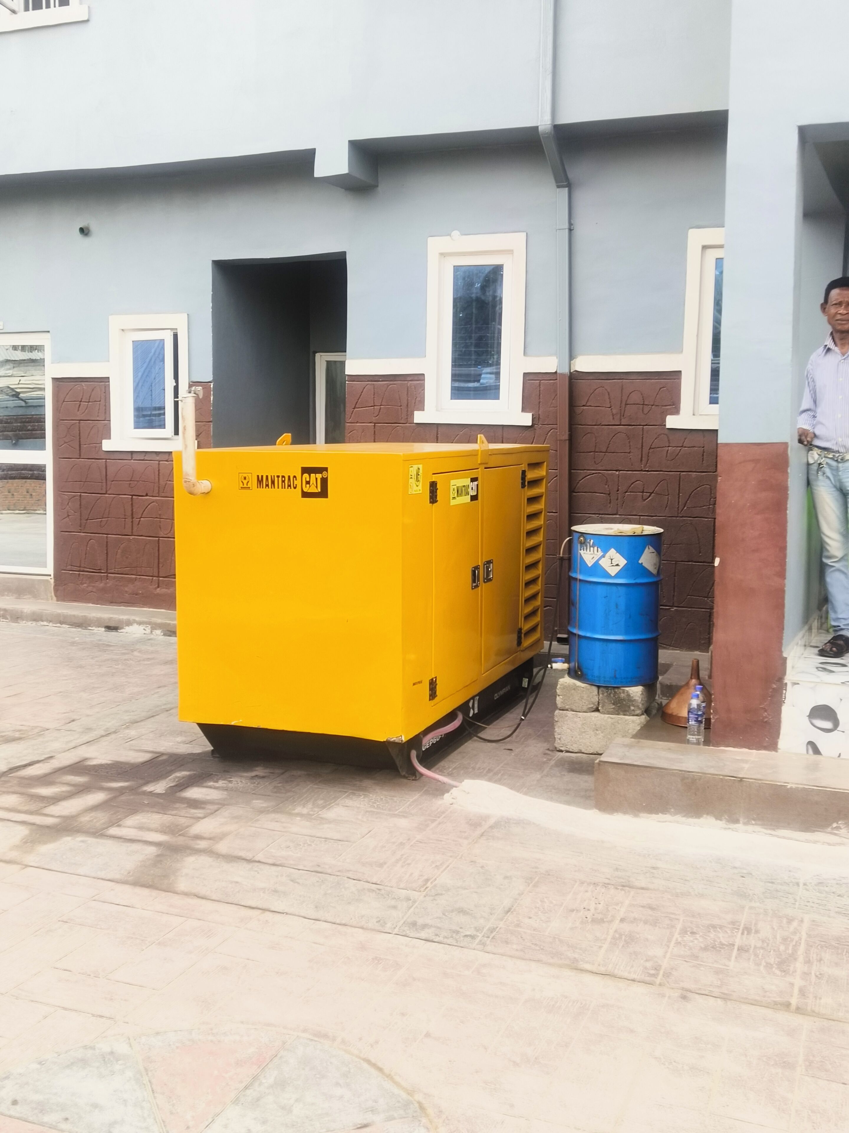 Een gele draagbare generator met de tekst 'MANTRAC CAT' geparkeerd voor een gebouw met een raam en een deur.