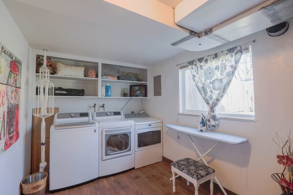 Una sala de lavandería con lavadora, secadora, tabla de planchar y estantes con diversos objetos.