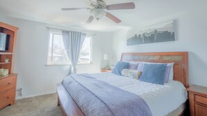 Une chambre à coucher comprenant un lit, une tête de lit en bois, des tables de chevet, un ventilateur de plafond, une fenêtre avec des rideaux et une œuvre d’art représentant un paysage urbain accrochée au mur.