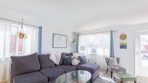 Un salon avec un canapé de couleur sombre, une table basse en verre, un fauteuil et une petite table avec une lampe. Il y a des rideaux aux fenêtres, une plante suspendue et une carte encadrée au mur.
