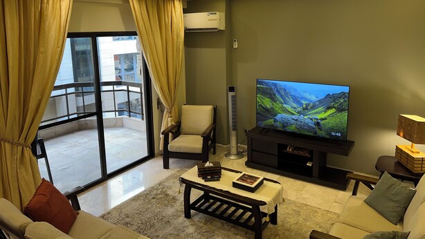 Sala de estar com televisão de tela plana, sofá, mesa de centro e uma poltrona. Há uma varanda com porta de vidro e cortinas.