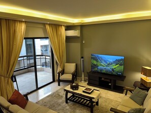 Un salon équipé d’un téléviseur à écran plat, d’un canapé, d’une table basse et d’un fauteuil. Il y a un balcon avec une porte vitrée et des rideaux.