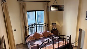 Quarto com cama, mesa de cabeceira com abajur, janela com cortinas e radiador.