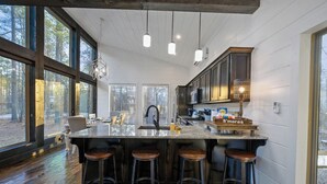 A modern kitchen with a large island, bar stools, and a view of the forest through large windows. - Luxury Farmhouse Cabin-3 King Suites + Bunk/Game (Broken Bow)