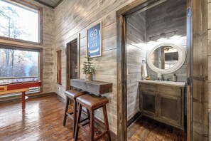 A rustic bathroom with wooden walls, a wooden bench, a sign that says "GO JUMP IN THE LAKE", and a round mirror. - Luxury Farmhouse Cabin-3 King Suites + Bunk/Game (Broken Bow)