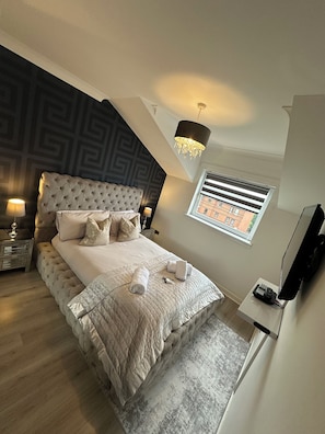 A bedroom with a tufted headboard, a bed with a quilted bedspread, two bedside lamps, a window with blinds, a flat-screen TV, and a telephone on a stand.