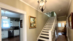 A staircase leading to a second floor, a hallway with a rug, and a kitchen area with cabinets and a chandelier. - Aggie House near ASU (Jonesboro)
