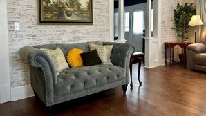 A living room with a grey tufted sofa, a framed landscape painting, and a brick-patterned wall. - Aggie House near ASU (Jonesboro)