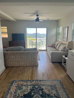 Ein Wohnzimmer mit einer Couch, einem Couchtisch, einem Fernseher, einem Deckenventilator und Blick auf Berge durch eine Schiebetür.