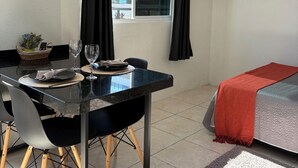 A hotel room with a dining table, chairs, a bed with a red blanket, a TV mounted on the wall, and a window with curtains. - Loft for couples - Downtown Bombinhas - Near the beach (Bombinhas)