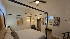 Uma cama com dossel, cabeceira, ventilador de teto, ar condicionado, um quadro, uma cadeira e uma porta que dá para outro quarto.