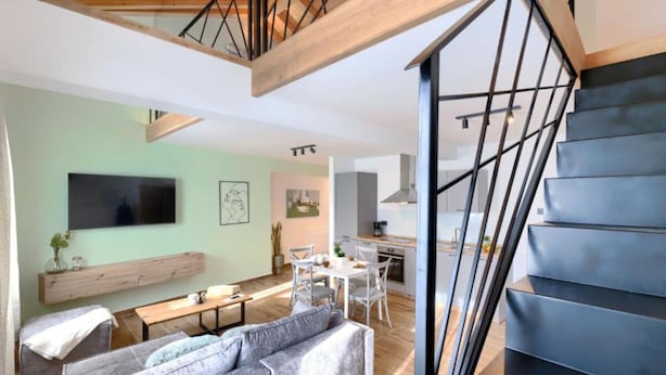 Ein modernes Wohnzimmer mit einer Couch, einem Couchtisch und einem Essbereich mit Stühlen und Tisch. Eine Treppe mit Metallgeländer führt auf eine obere Ebene.