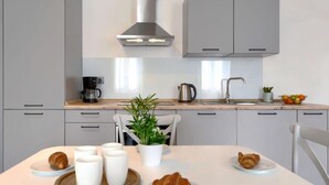 Eine moderne Küche mit grauen Schränken, einer Edelstahl-Dunstabzugshaube, einer Kaffeemaschine, einem Wasserkocher und einer Pflanze auf der Arbeitsfläche. Auf dem Tisch stehen Croissants auf Tellern.