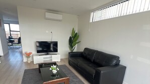 A modern living room with a black leather sofa, a wooden coffee table with flowers, a flat-screen TV, and a large plant. - SJ House easy access to the Airport and two minutes walking distance to the Mall (Takanini)