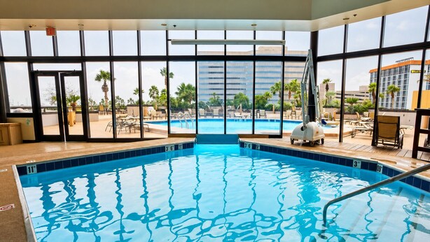 An indoor swimming pool with clear glass walls, a lifeguard chair, and a view of palm trees and buildings outside.