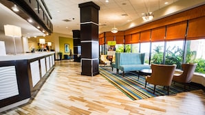 A hotel lobby with a reception desk, seating area with chairs and a sofa, a colorful striped rug, and large windows with a view of greenery.
