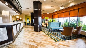 Eine Hotelhalle mit einer Rezeption, einer Sitzecke mit Stühlen und einer Couch, einem farbenfrohen StreifenTeppich und großen Fenstern mit Blick auf Grünflächen.