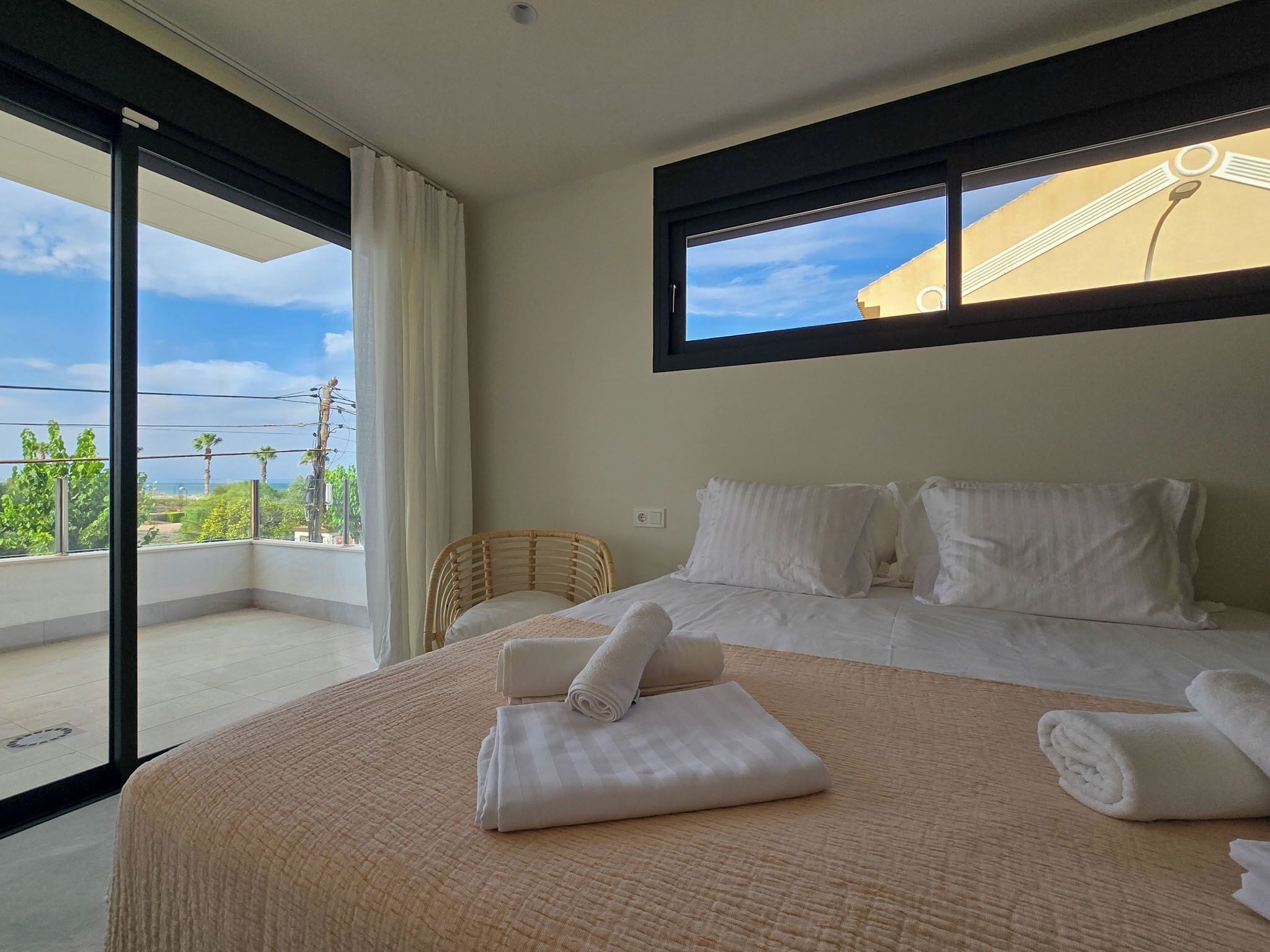 A hotel room with a large bed, a balcony with a view of palm trees and the sea, and a window with a view of a building.