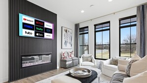 A modern living room with a flat-screen TV displaying various streaming services, a fireplace, and a large window offering a view of the outdoors. - Upscale 3BR Townhome – Walk to Shops & The Monon! (Carmel)