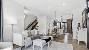 A modern living room with a sofa, ottoman, and coffee table, a staircase, and a dining area with a chandelier. - Upscale 3BR Townhome – Walk to Shops & The Monon! (Carmel)
