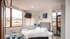A modern bedroom with a large bed, a TV mounted on the wall, a dining area with a table and chairs, and a balcony with a view of the city. - Cozy Suite in the Heart of Historic Cuenca awaits you (Cuenca)