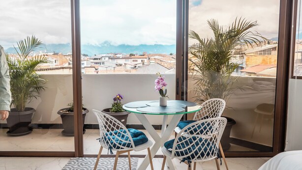 A balcony with a glass door, two potted plants, a round table with chairs, and a view of buildings and mountains. - Cozy Suite in the Heart of Historic Cuenca awaits you (Cuenca)