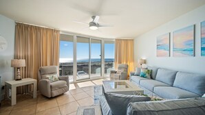 Een ruime woonkamer met balkon, plafondventilator en een groot raam met gordijnen.