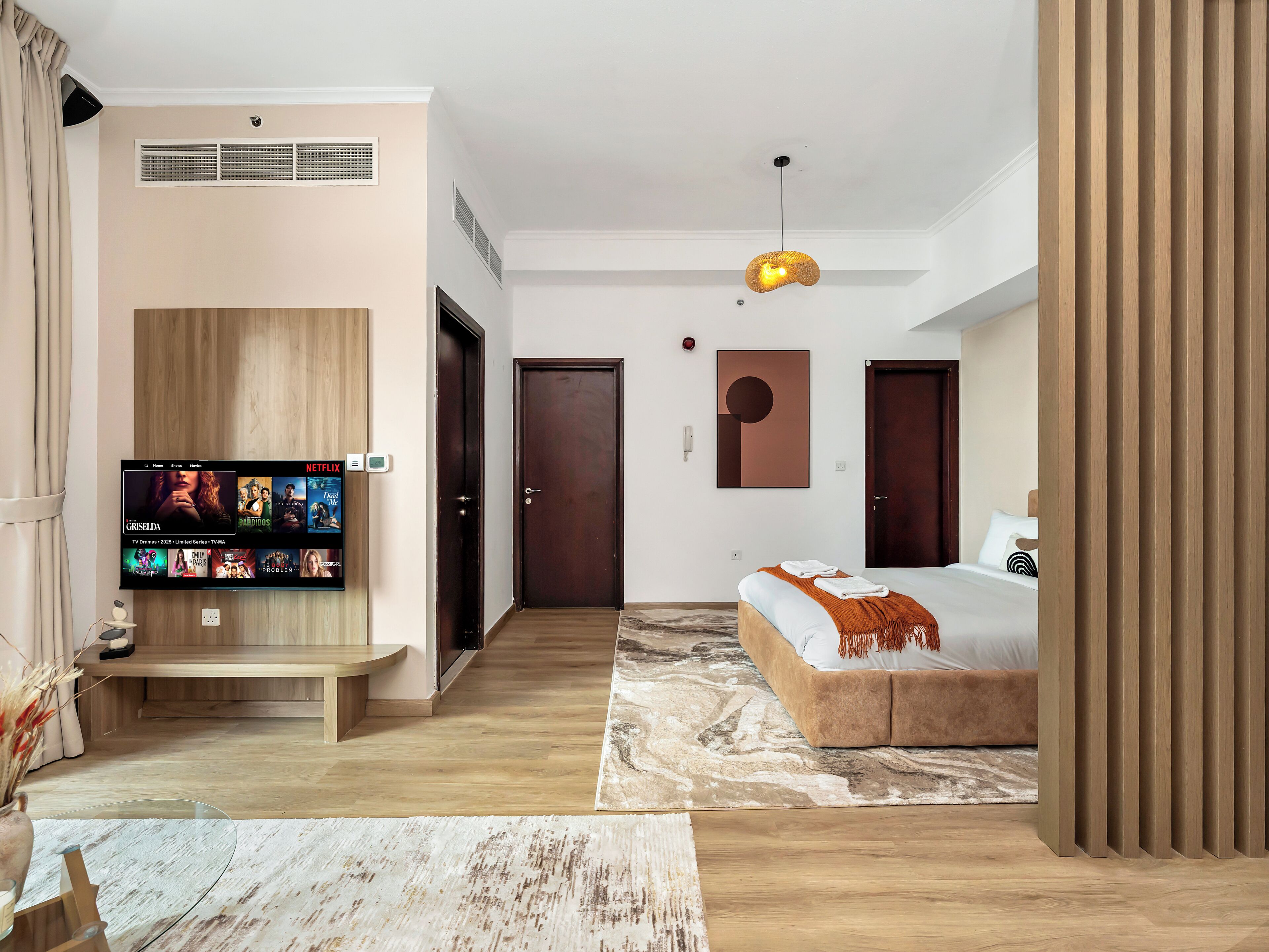 Una habitación de hotel moderna con un televisor de pantalla plana grande, una cama con cabecero marrón, un banco de madera y una obra de arte original en la pared.