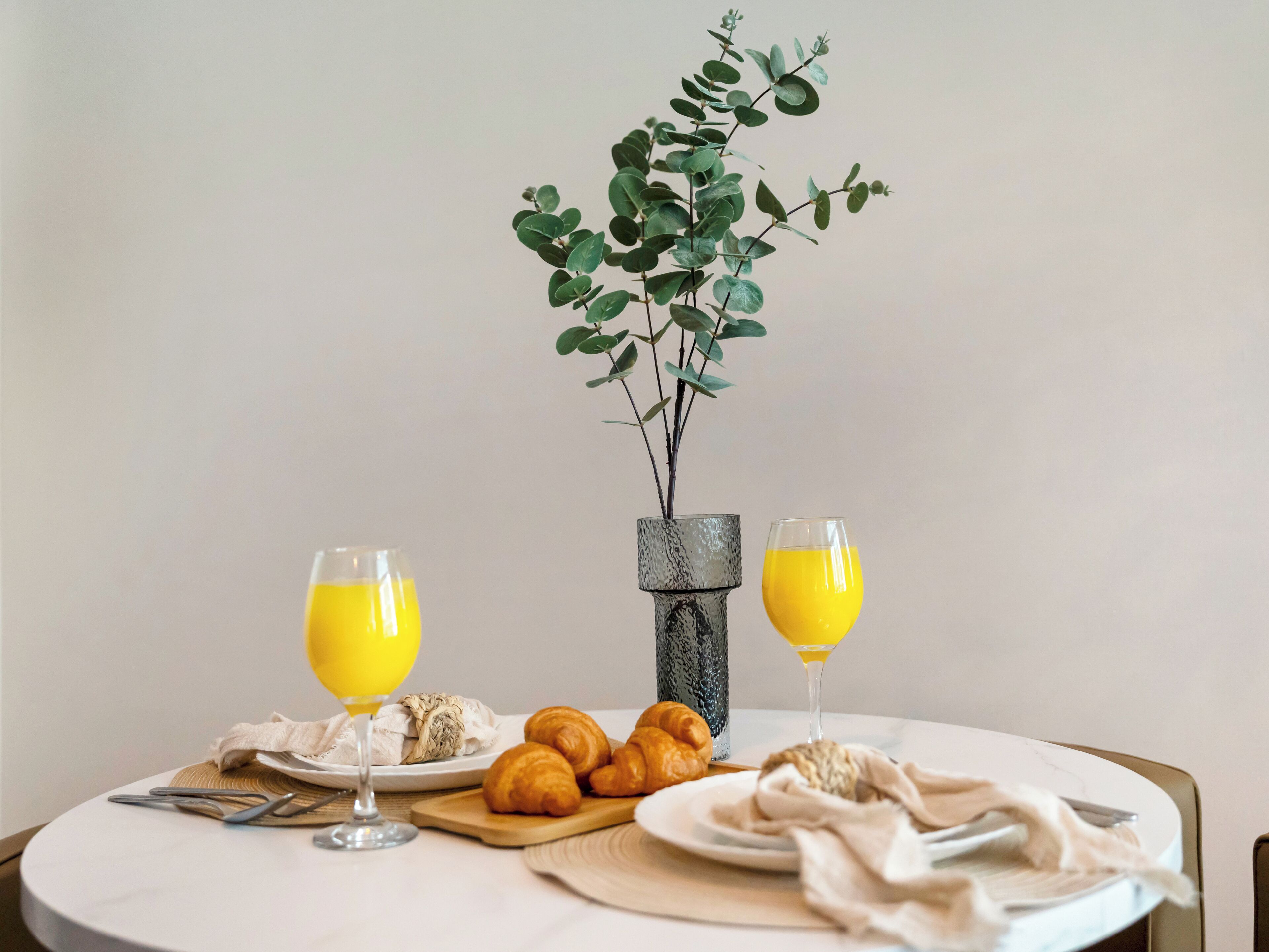 Una mesa de desayuno con croissants, un florero con una planta y dos vasos de jugo de naranja.