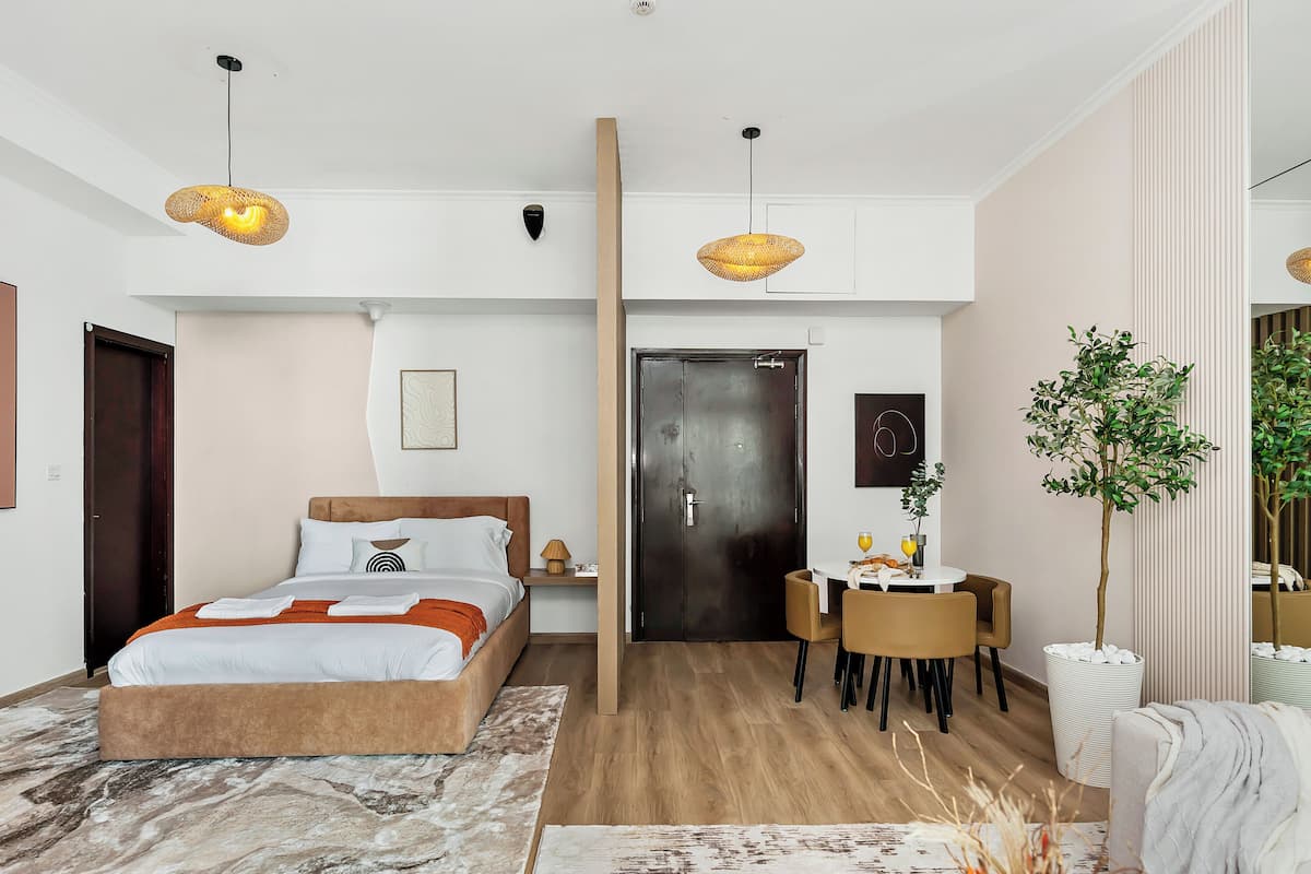 A modern hotel room with a bed, bedside table, two hanging lamps, a dining area with chairs, a small table, and a potted plant.