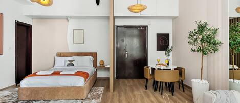 A modern hotel room with a bed, bedside table, two hanging lamps, a dining area with chairs, a small table, and a potted plant.