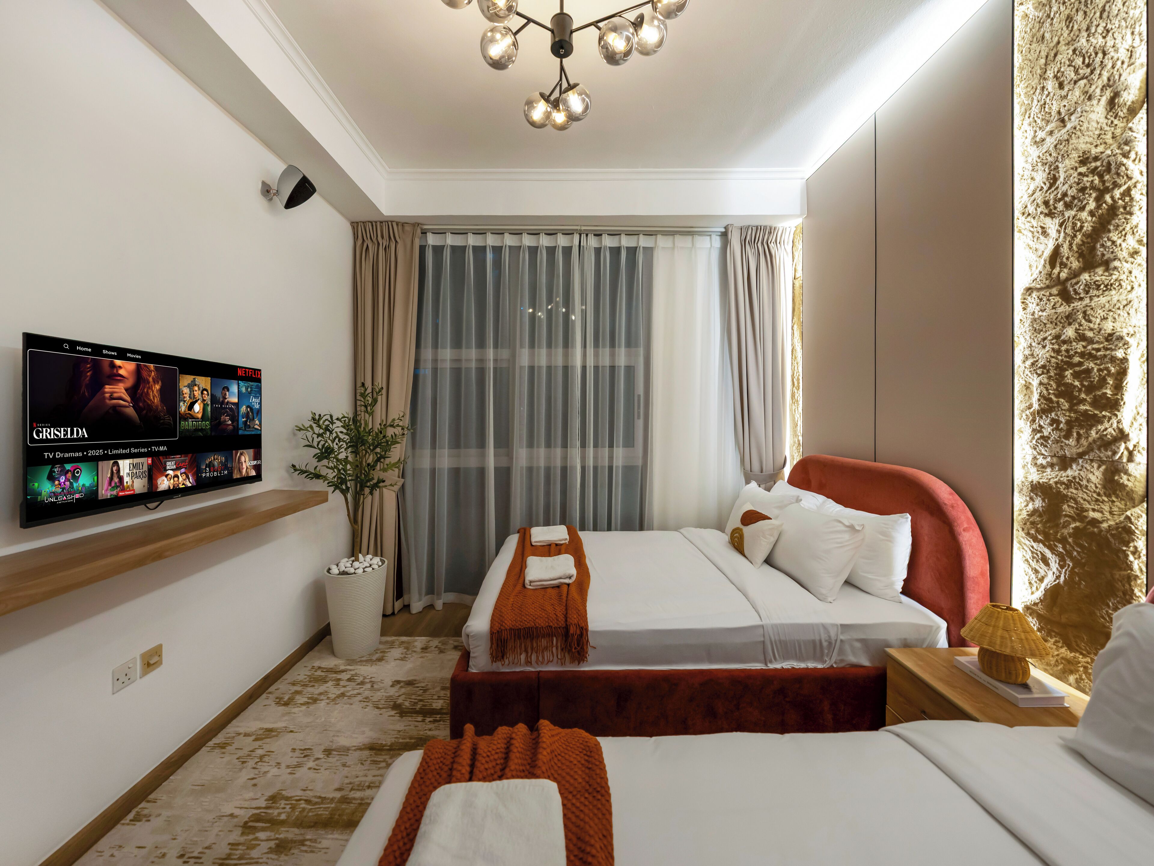 A modern hotel room with a large flat-screen TV displaying a movie selection, a bed with white linens and a red headboard, a small table with a lamp, and a large window with sheer curtains.