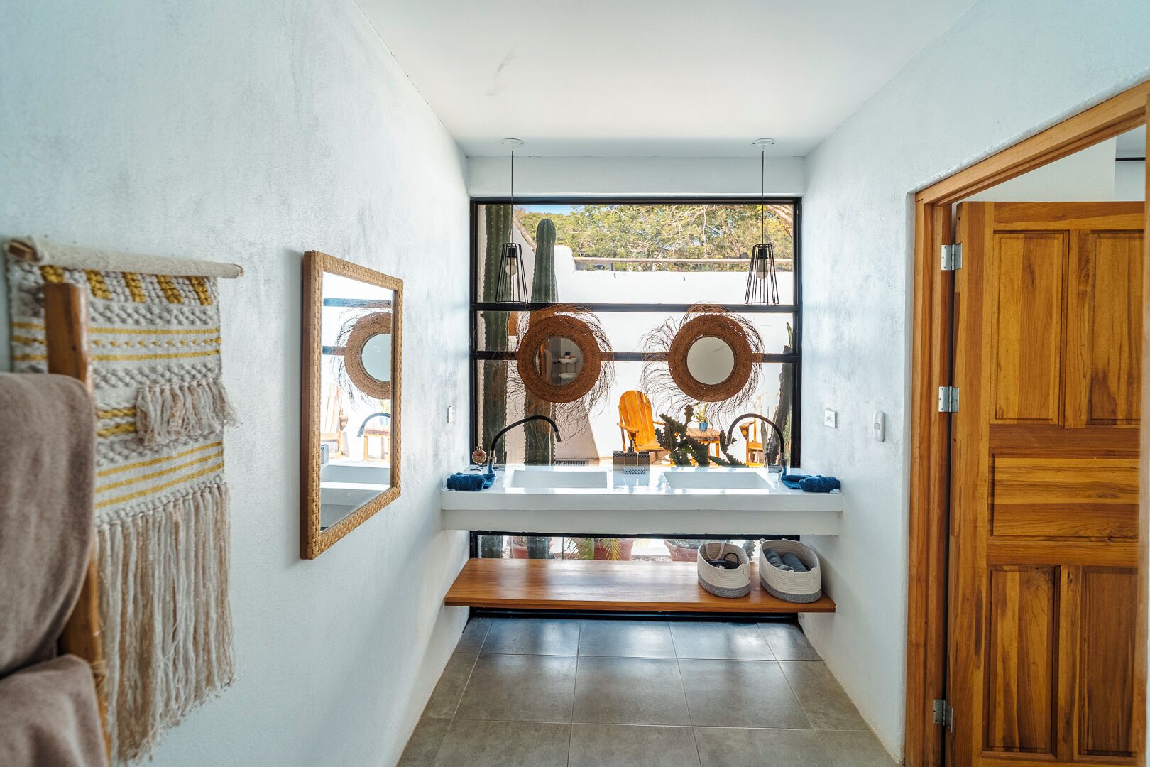A narrow hallway with a white wall, a wooden door, and a window with a view of a cactus and outdoor furniture.