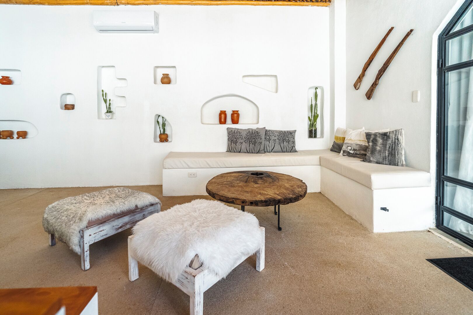 A modern living room with a white sofa, two white furry ottomans, a round wooden coffee table, and a glass door leading outside.