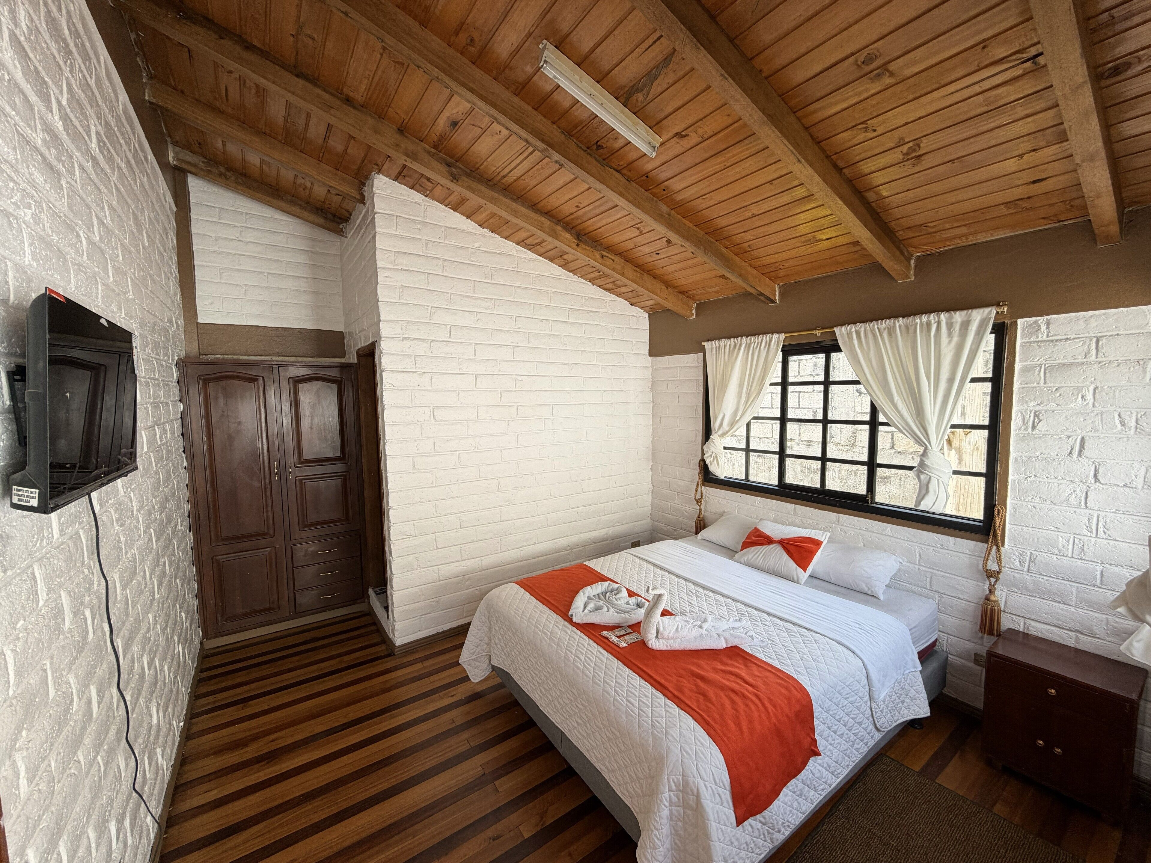 Une chambre avec un plafond en bois, des murs en briques blanches, un lit avec une literie blanche et orange, une armoire en bois et une fenêtre avec des rideaux.