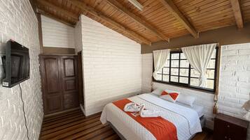 Une chambre avec un plafond en bois, des murs en briques blanches, un lit avec une literie blanche et orange, une armoire en bois et une fenêtre avec des rideaux.