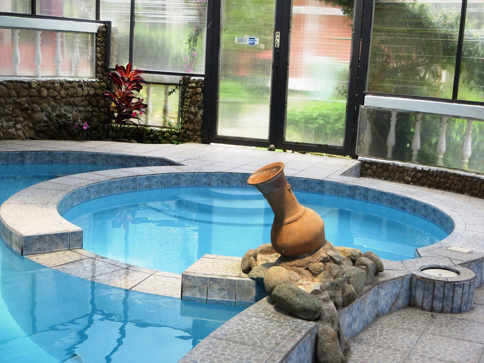 A swimming pool with a unique terracotta vase centerpiece, surrounded by stone tiles and a stone wall.