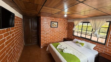 Una habitación con pared de ladrillo, cama con ropa de cama verde y blanca, techo de madera, televisor montado en la pared y una ventana con cortinas.
