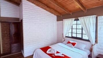 A room with a wooden ceiling, white brick walls, a bed with red bedding, and a window with white curtains.